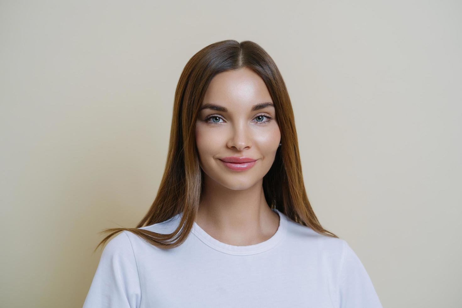 tierna mujer femenina mira con admiración y afecto, usa camiseta blanca, tiene maquillaje, piel sana, cabello oscuro, mira algo maravilloso, aislado sobre la pared beige del estudio. belleza, cosmetologia foto