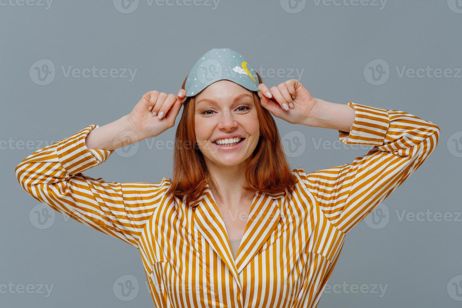 Positive redhaired young female wears eye mask for sleep on head, smiles gladfully, dressed in home wear, has sweet dreams at night, isolated over grey background, enjoys peaceful atmosphere photo