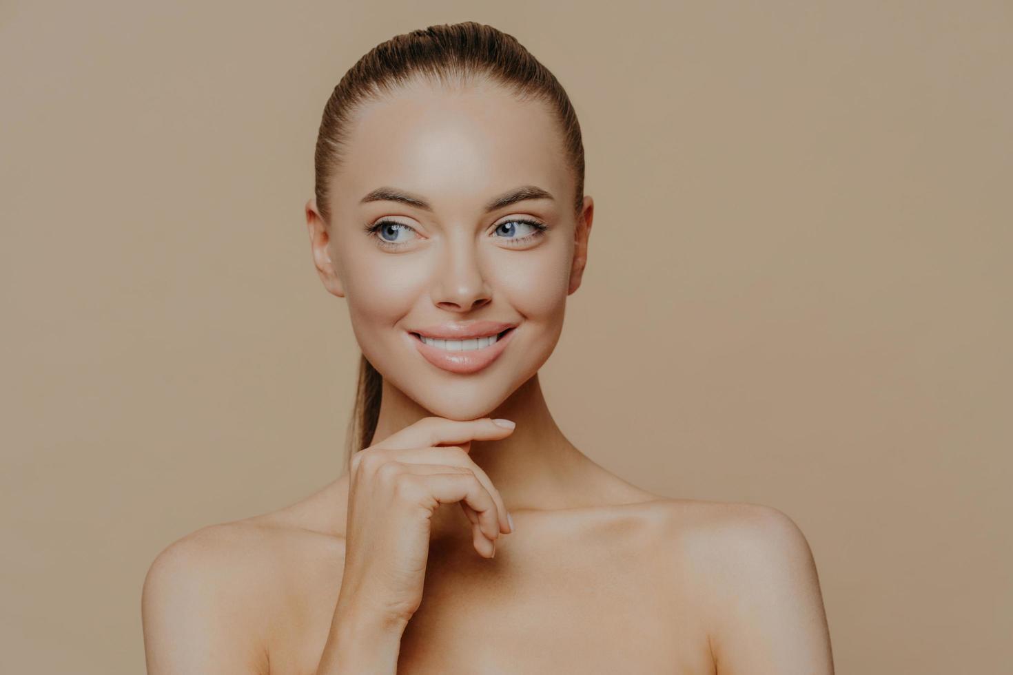 mujer joven sonriente y bien arreglada con cuidado facial de belleza, piel brillante y saludable, toca la barbilla suavemente y mira a un lado, posa sin camisa contra el fondo marrón del estudio, ama su piel, se siente renovada foto