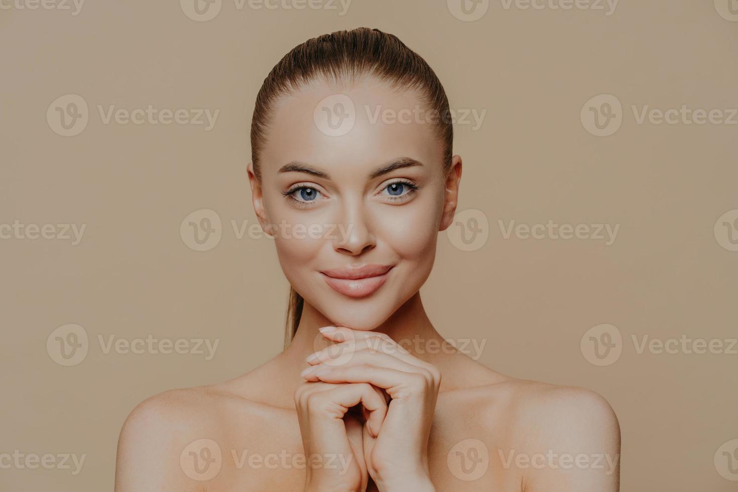 Young pretty woman with beauty skin and combed hair, keeps hands under chin, has healthy complexion after face purification, soft well cared body, isolated over studio wall. Facial treatment photo
