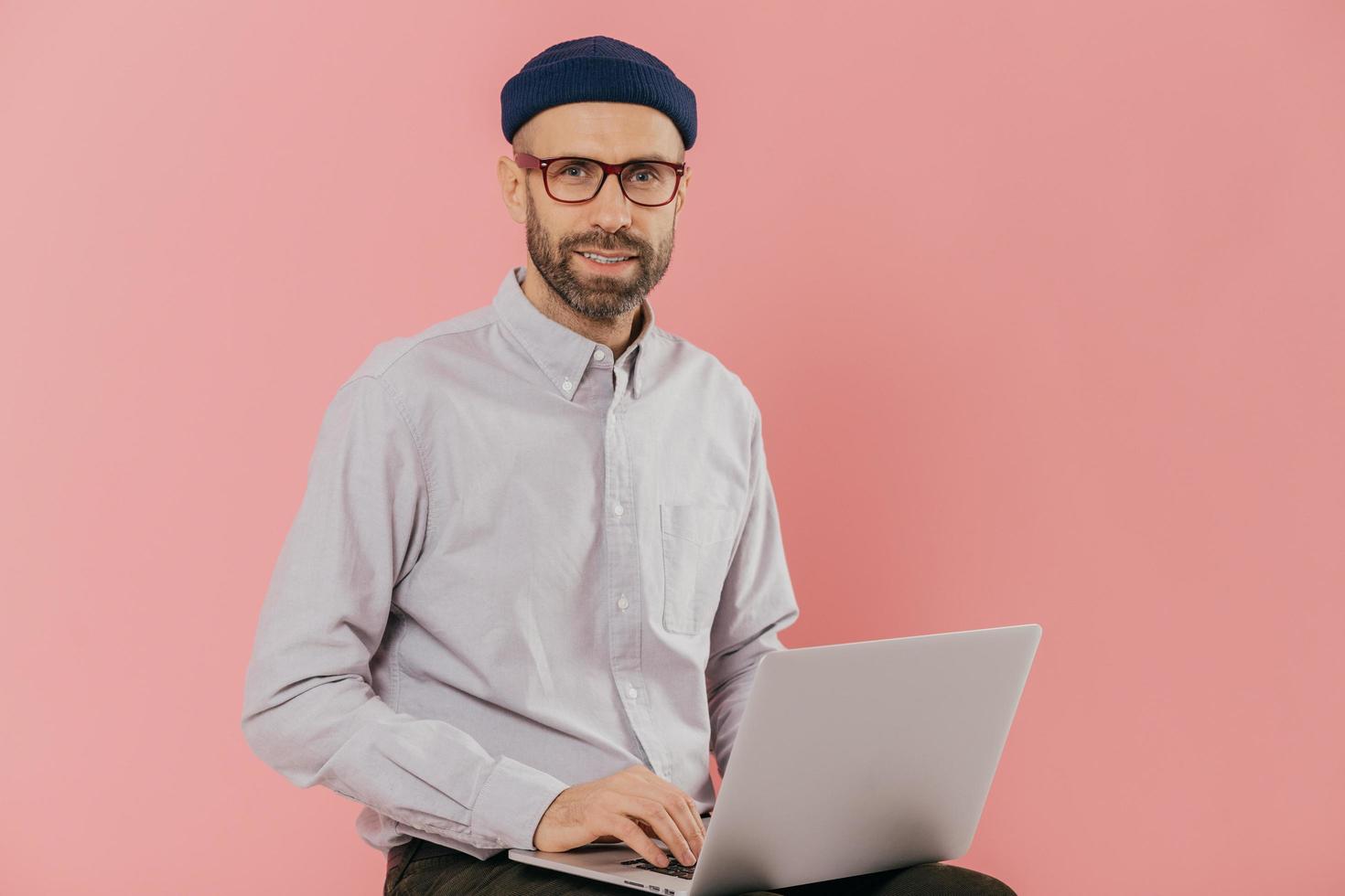 Male graphic designer thinks about creative ideas, reads news on internet website, uses moderm laptop computer, sits against pink background. Businessman reads publication online, makes research photo