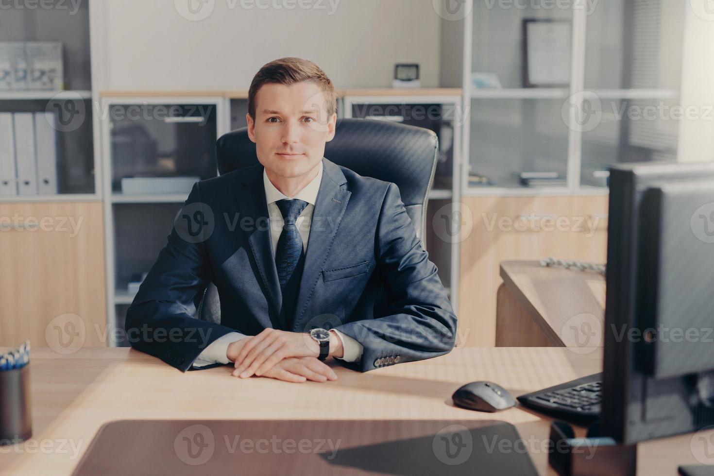 Proud handsome male entrepreneur sits in his cozy cabinet, works on computer, satisfied with successful working day, dressed formally, looks directly into camera. Intelligent employee in office photo