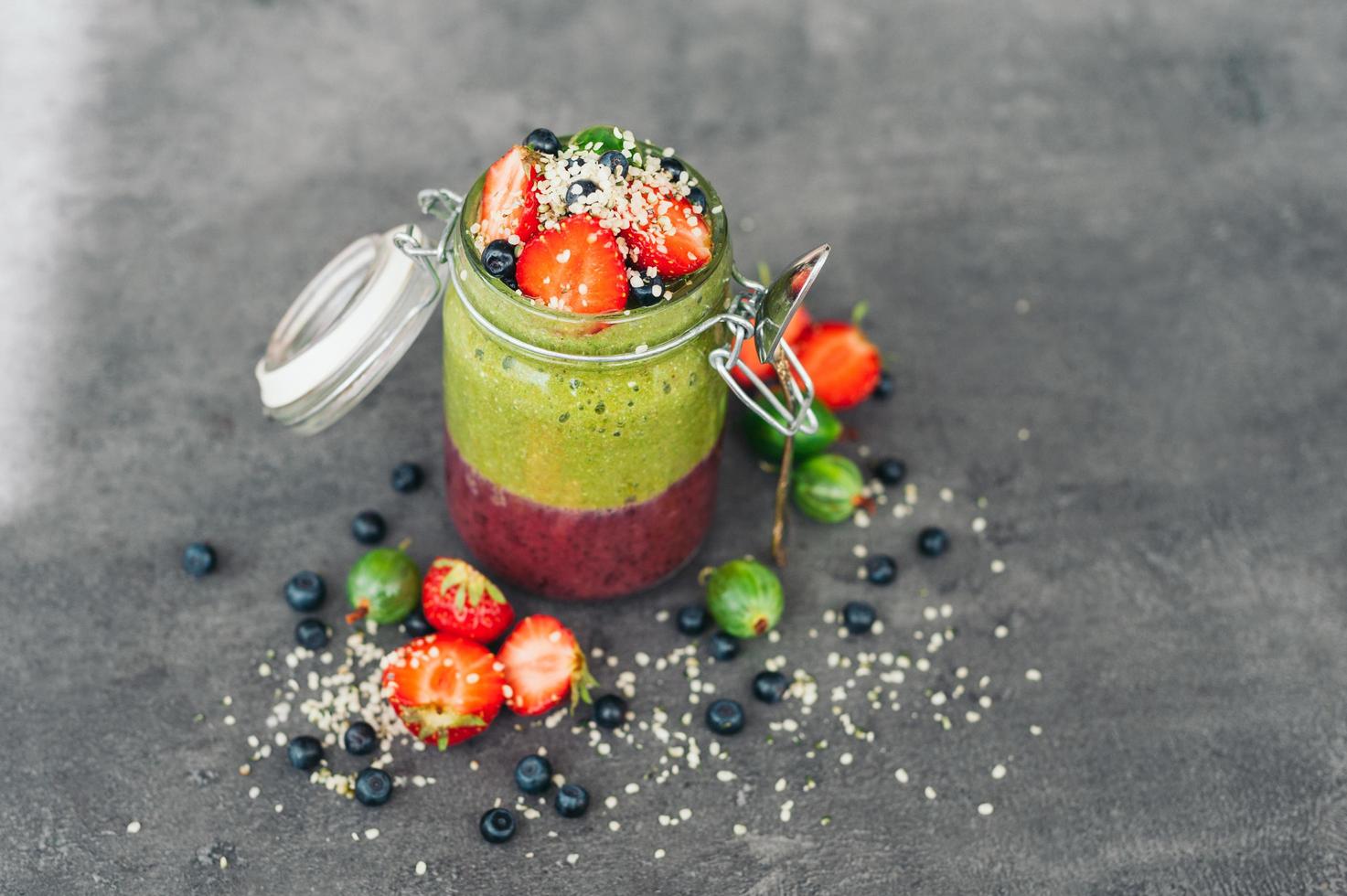 tarro de batido de frutas con frutas exóticas fresa, grosella y arándano con copos de coco aislado sobre fondo gris. postre delicioso foto