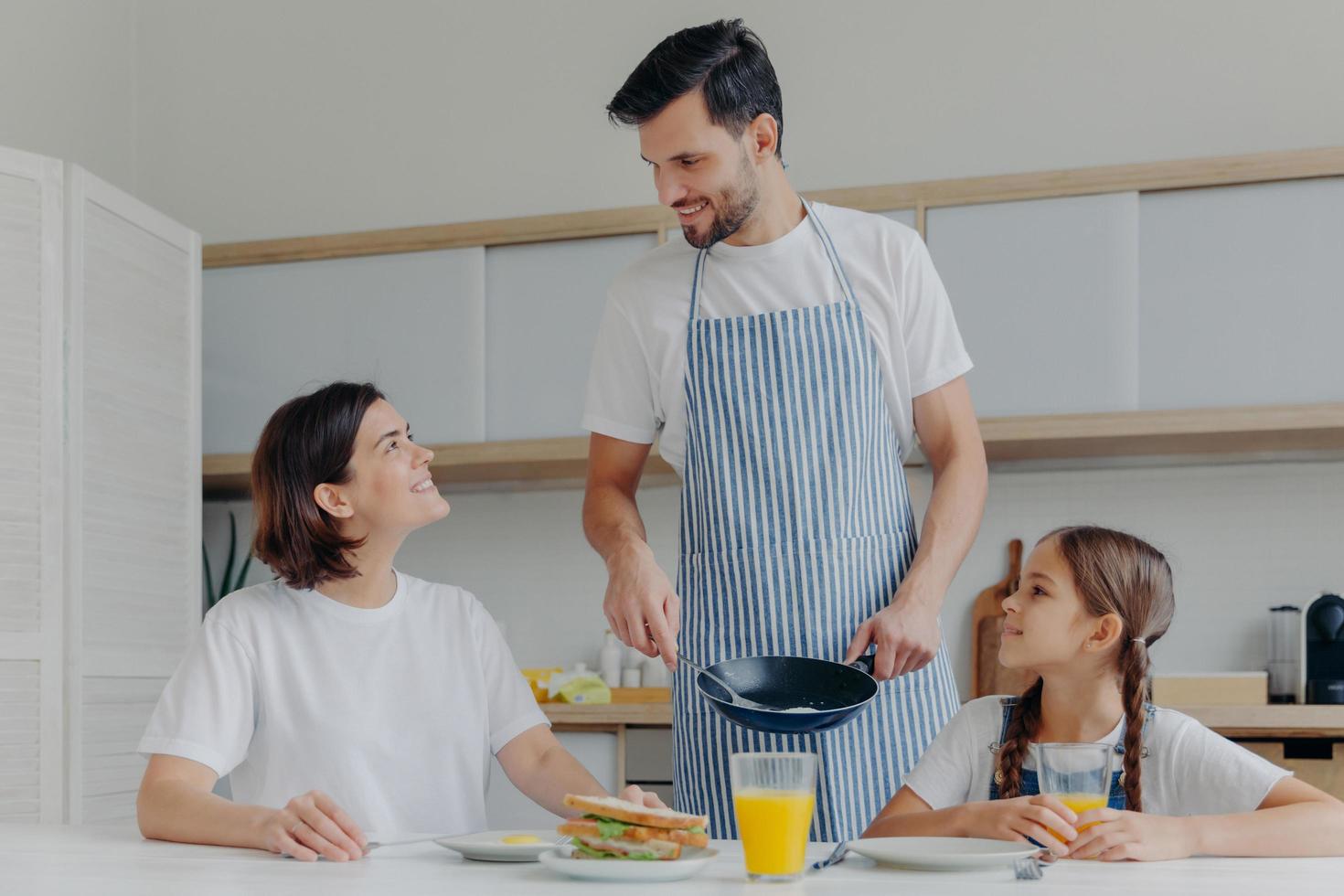 Happy father, mother and daughter gather together at kitchen, eat delicious breakfast, dad prepared fried eggs, being in good mood, ready to start new day. Lovely family enjoy tasty meal at home photo