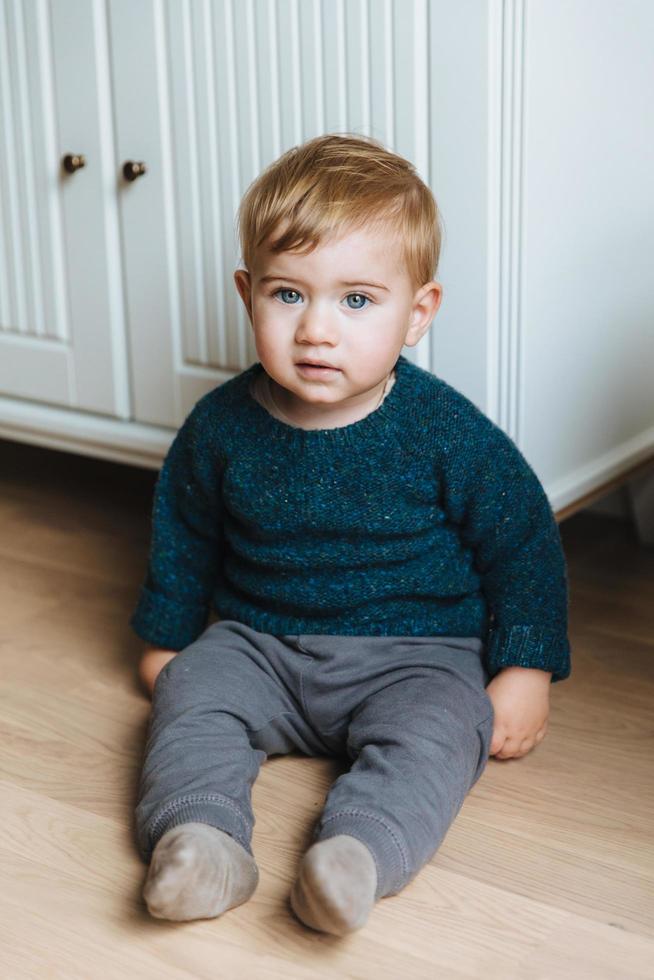 concepto de niños. pequeño bebé pequeño con atractivos ojos azules, mejillas regordetas y cabello rubio se sienta en el piso, el maestro va solo, usa suéter y pantalones. niño pequeño con expresión seria foto