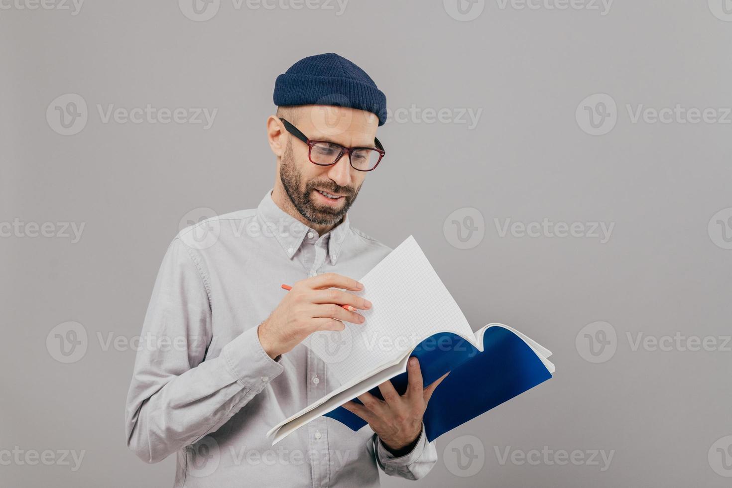 Photo of handsome unshaven young guy holds notebook, turns over page, writes records and planning for next week, wears optical glasses and white formal shirt, models over grey background alone