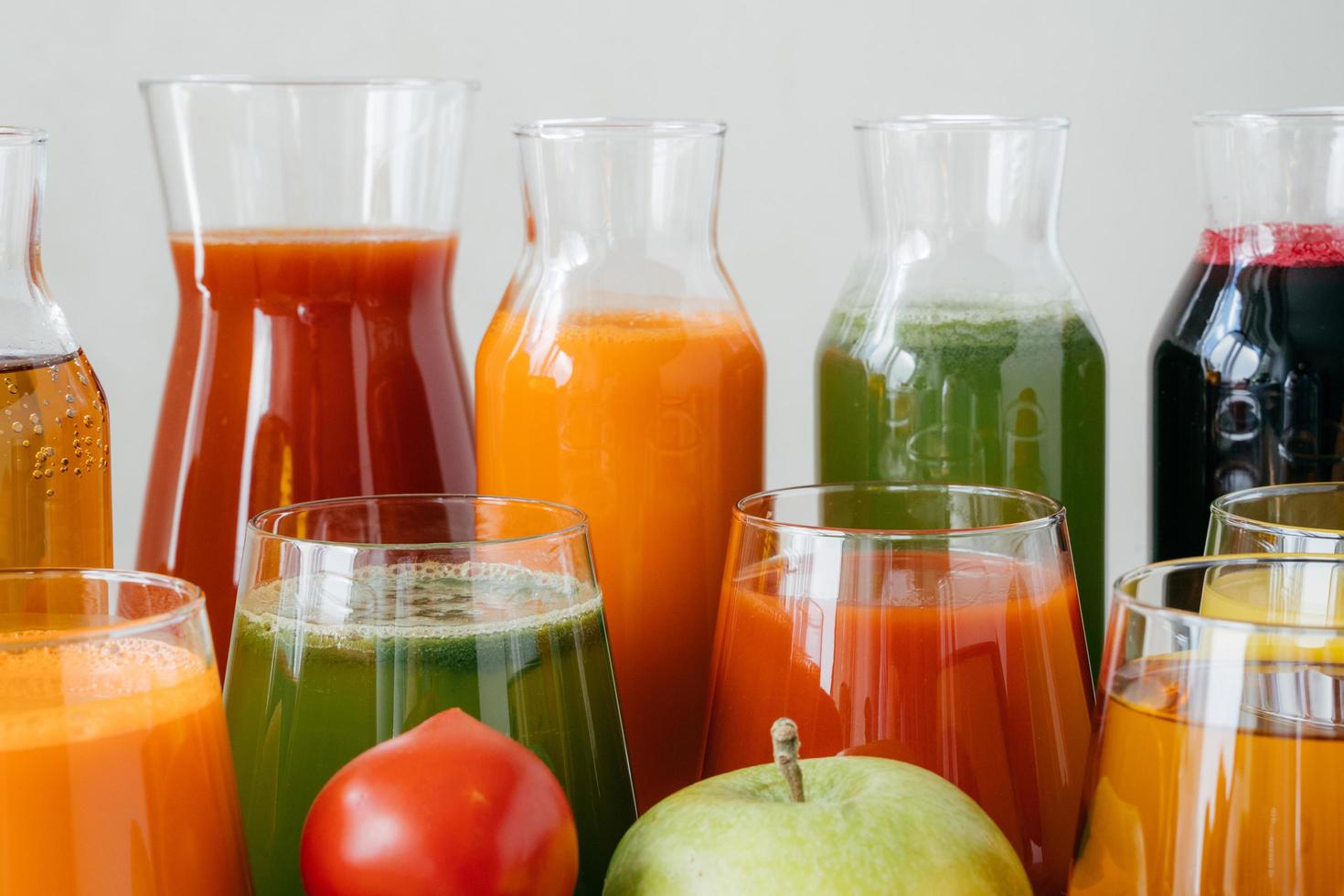 Close up shot of glass bottles filled with colorful juice made of various vegetables and fruit, red tomato and green apple in foreground. Fresh detox drink photo
