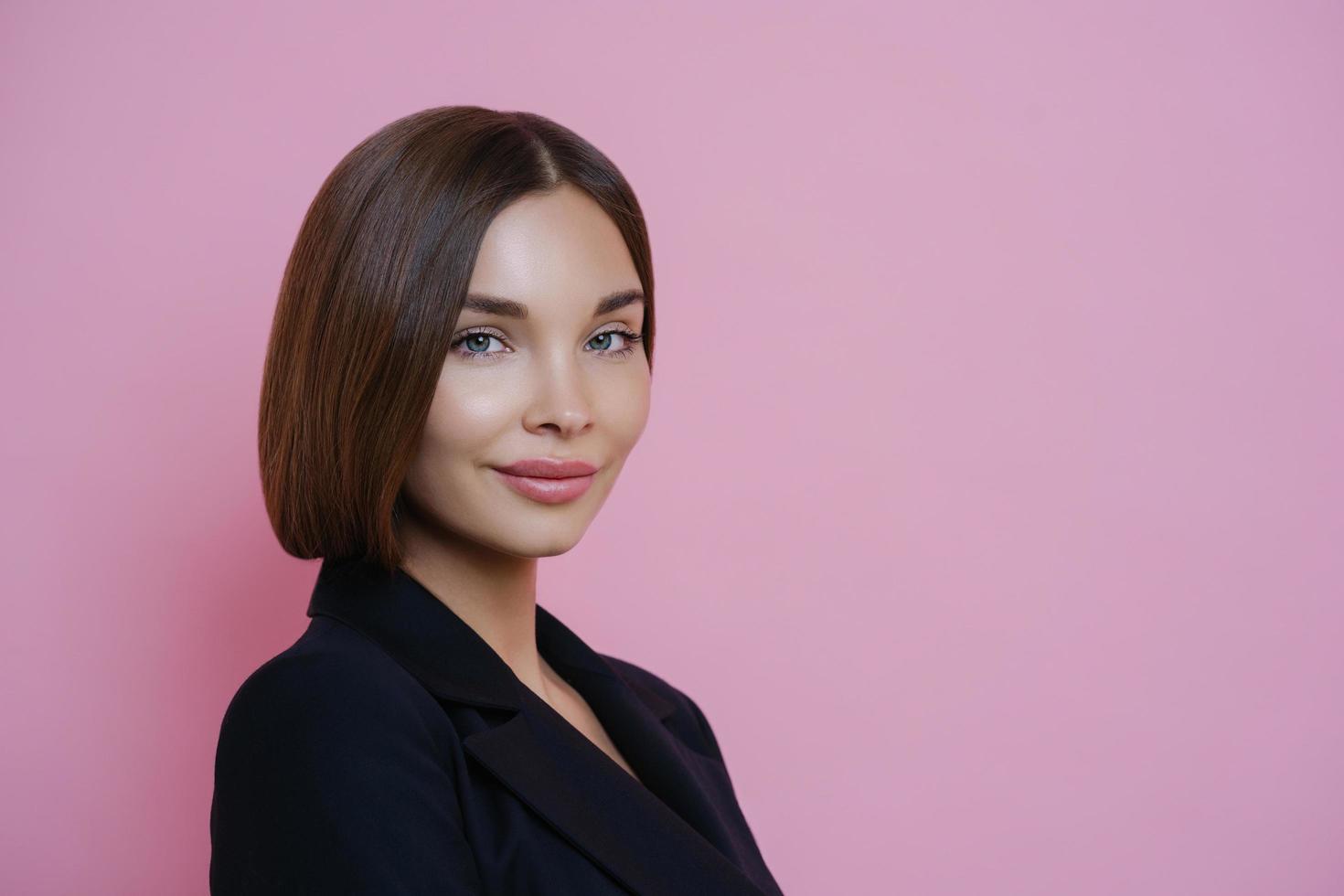 foto de perfil de una mujer de cabello oscuro vestida con traje negro, tiene maquillaje natural, se ve segura de sí misma en el interior, posa sobre fondo rosa, espacio vacío para su promoción. concepto de mujer y elegancia