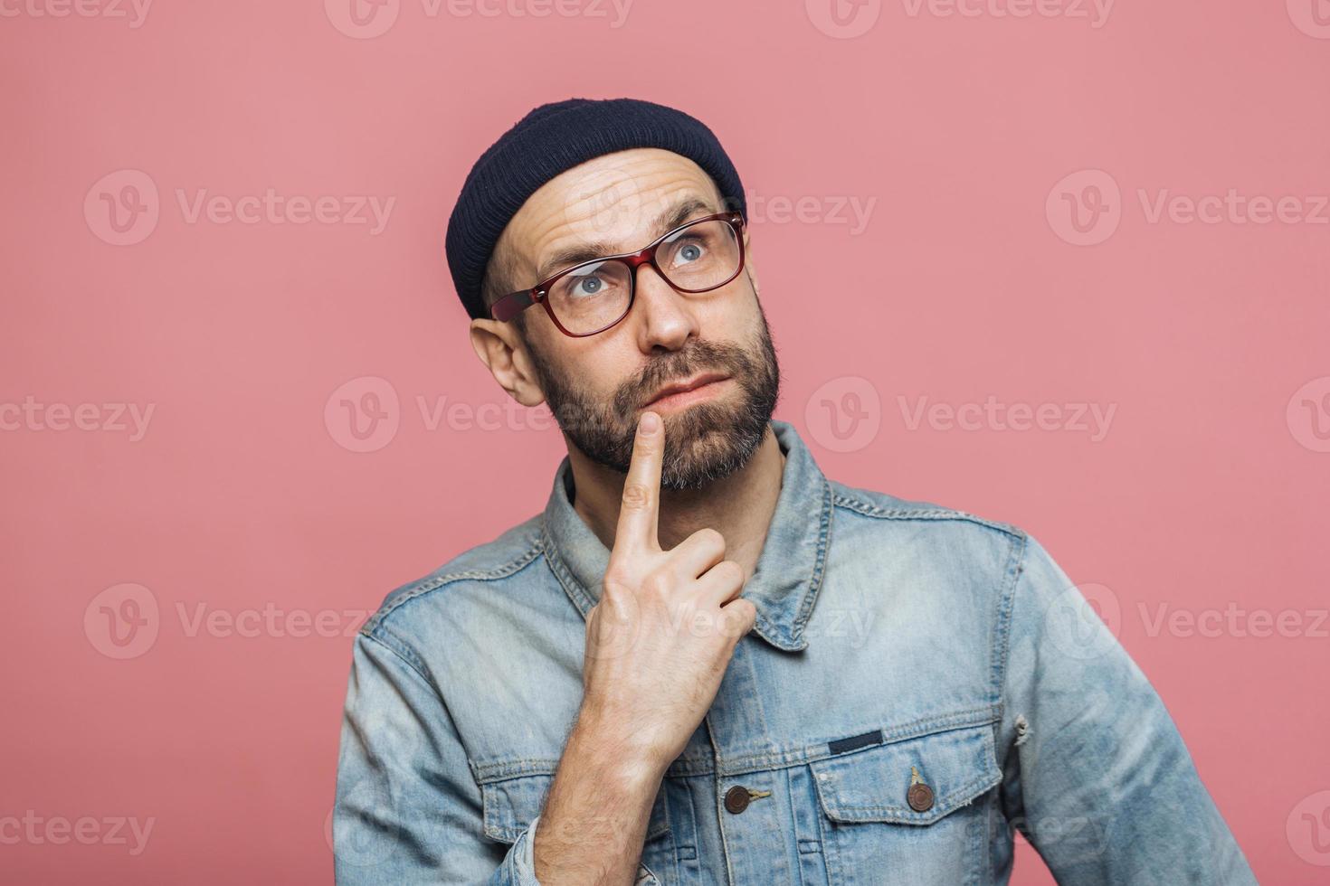 Thoughtful bearded male being deep in thoughts, looks with pensive expression into camera, concentrated on something, poses against pink background. People, facial expressions, daydreaming concept photo