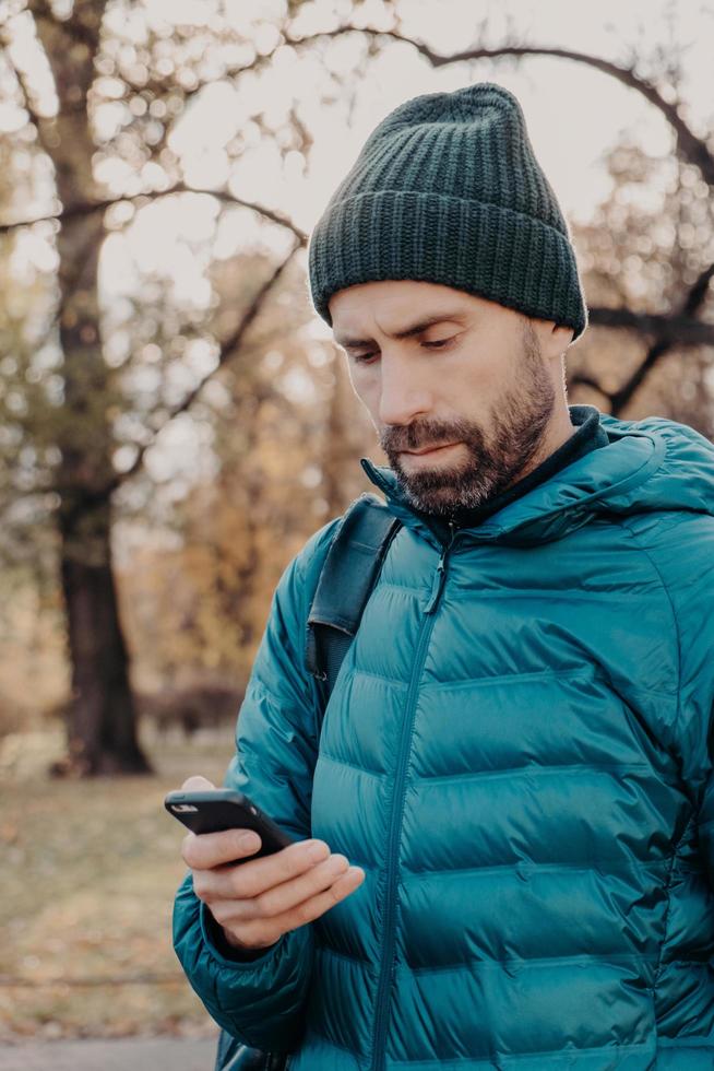 toma vertical de un tipo hipster concentrado con barba gruesa, usa anorak y sombrero, tiene celular, usa una aplicación especial para encontrar ruta, mensaje con un amigo, respira aire fresco. concepto de estilo de vida foto