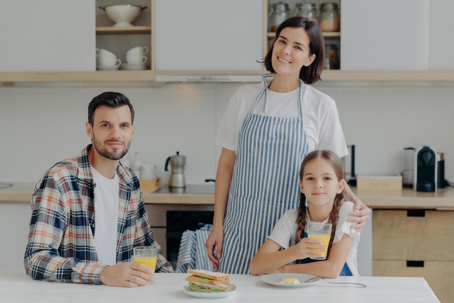 Family consists of mother, father and daughter pose together in modern kitchen, drink fresh juice, eats sadwiches, enjoy domestic atmosphere. Lovely woman in apron embraces small child with love photo