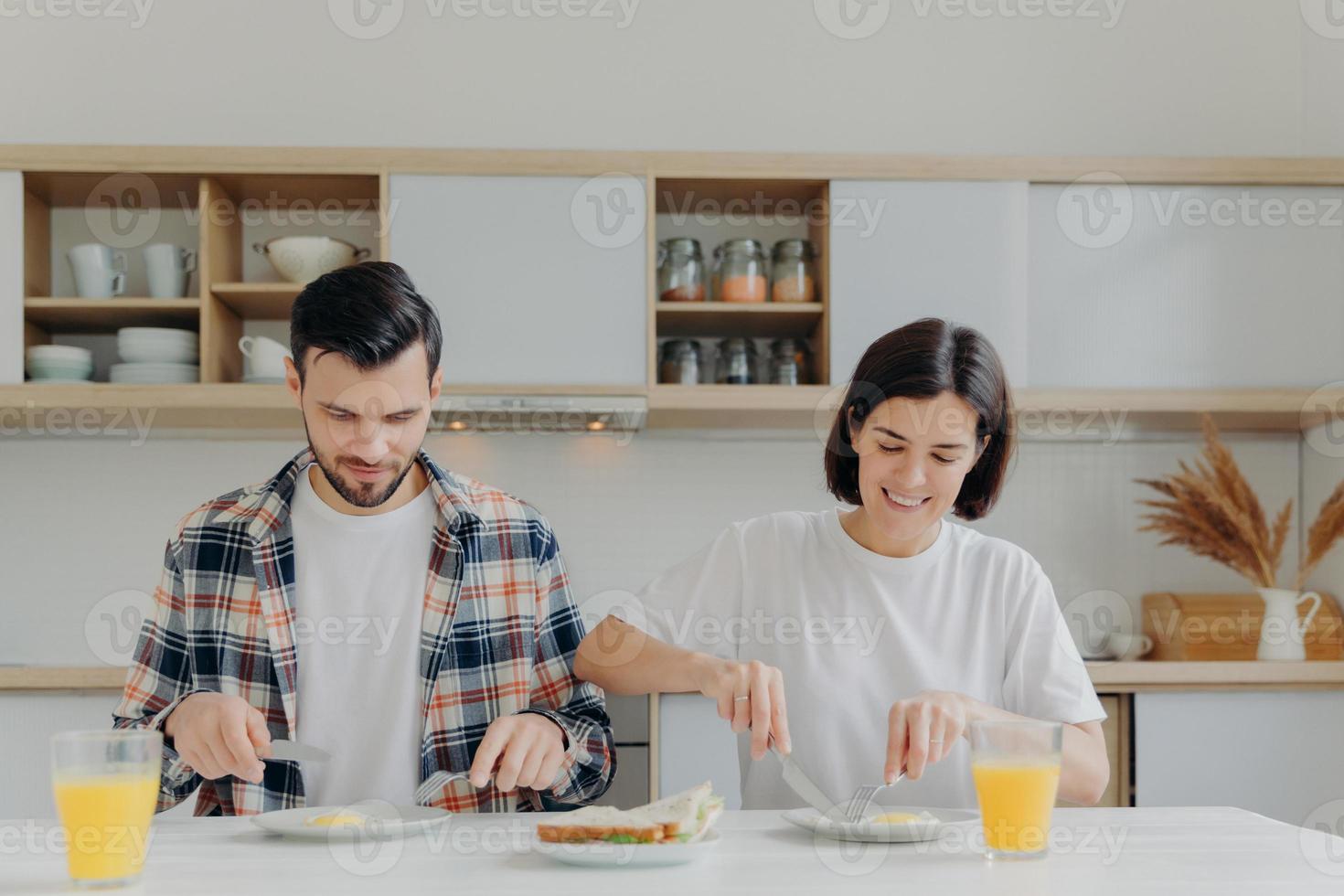 Family married couple pose at kitchen table, have delicious breakfast, talk about plannings on day, eat fried eggs and burgers, drink fresh apple juice, dressed casually, enjoy domestic atmosphere photo