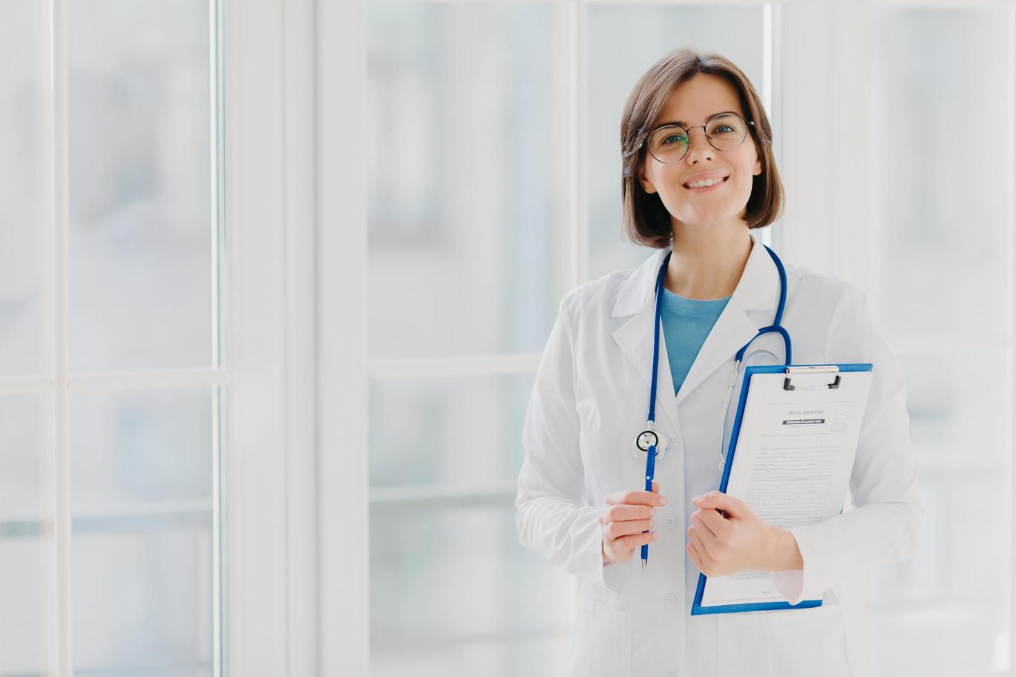 hermosa pasante sonriente sostiene portapapeles con papeles escritos y bolígrafo, usa uniforme blanco, lista para ver al paciente en su propia clínica privada, se encuentra en interiores contra fondo blanco. área de espacio de copia foto