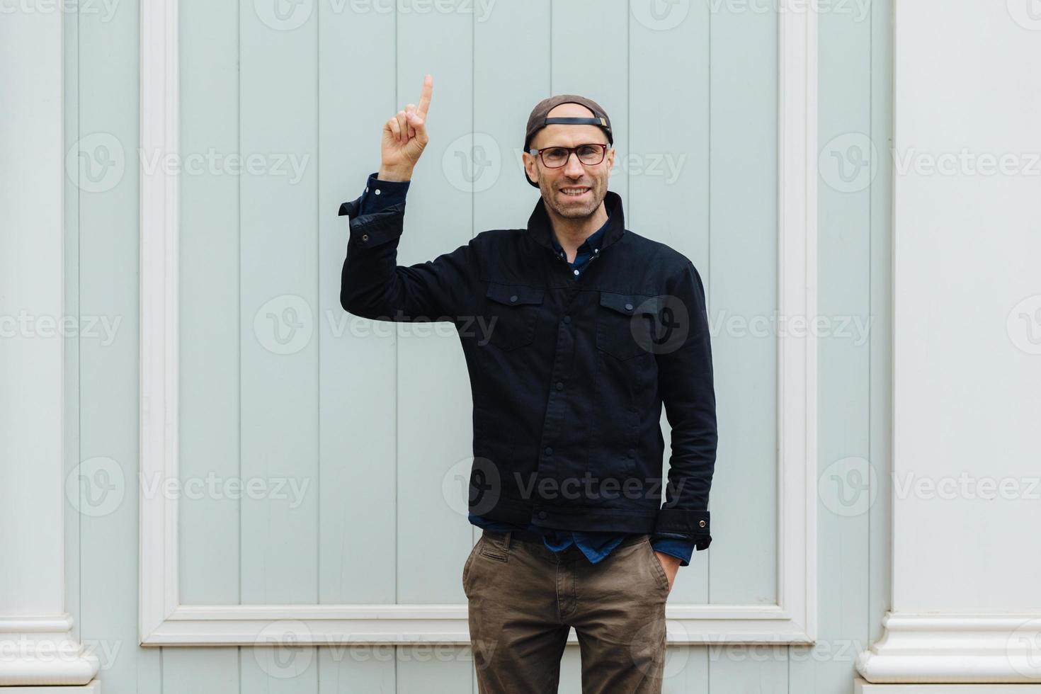 atractivo joven sin afeitar levanta el dedo índice, anuncia algo, mantiene la mano en el bolsillo, usa ropa elegante, posa solo contra la pared blanca, tiene una expresión encantada. oye, mira ahí foto