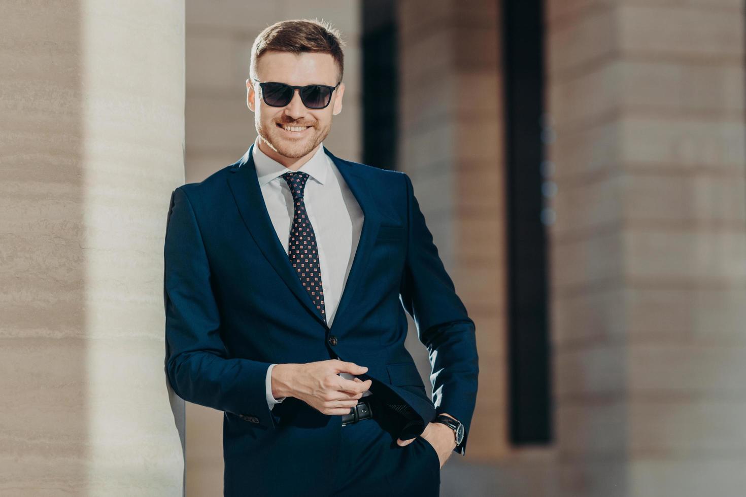 Horizontal shot of handsome cheerful bearded young male manager with confident cheerful expression, keeps hand in pocket, poses against office interior. People, business and elegance concept photo