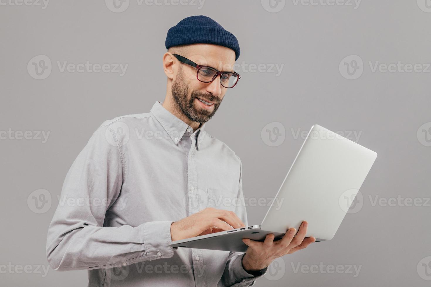 Shot of satisfied male with thick beard and mustache, holds laptop computer, transfers money, uses online banking service, sends files, connected to wireless internet, models over grey background photo