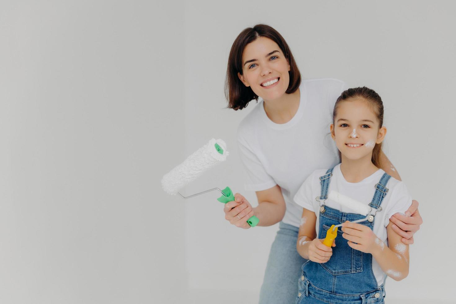 Family and house repairing concept. Positive caring mother cuddles child, have fun when refurbish walls, dressed in casual domestic clothes, hold repir tools, isolated over white background. photo