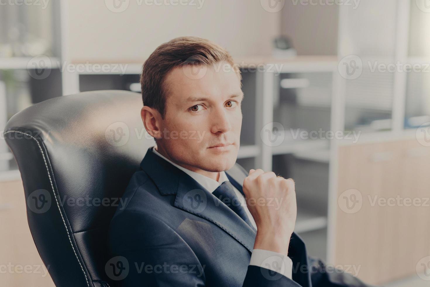 Indoor shot of serious confident businessman poses in chair, poses at cabinet, waits for colleagues to discuss main issues of work. Young male director in formal suit, thinks about new project photo