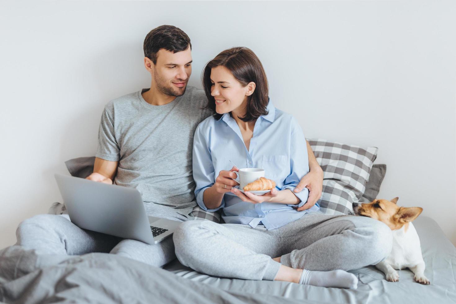 Affectionate couple in love embrace in bedroom, warch film online on laptop computer, have breakfast, enjoy domestic atmopshere and their favourite pet lies in bed. People, relatioships concept photo