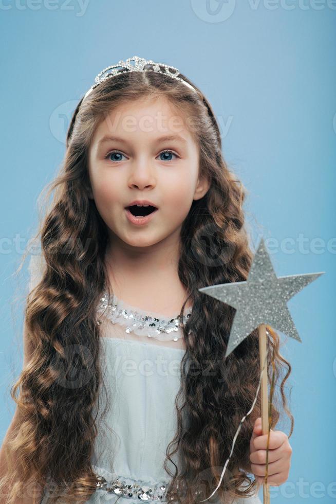 Small little child female princess wears crown and dress, holds magic wand, has long dark curly hair poses over blue background. Beautiful kid preapres for carnival or festive event. Childhood concept photo