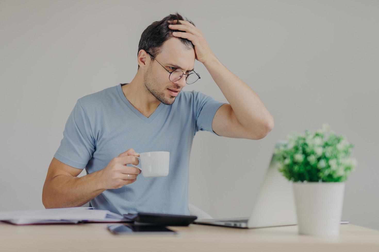 Young frustrated male does paperwork, reads with puzzled expression unpleasant information on laptop computer, drinks strong coffee, surrounded with calculator and documents on working table photo