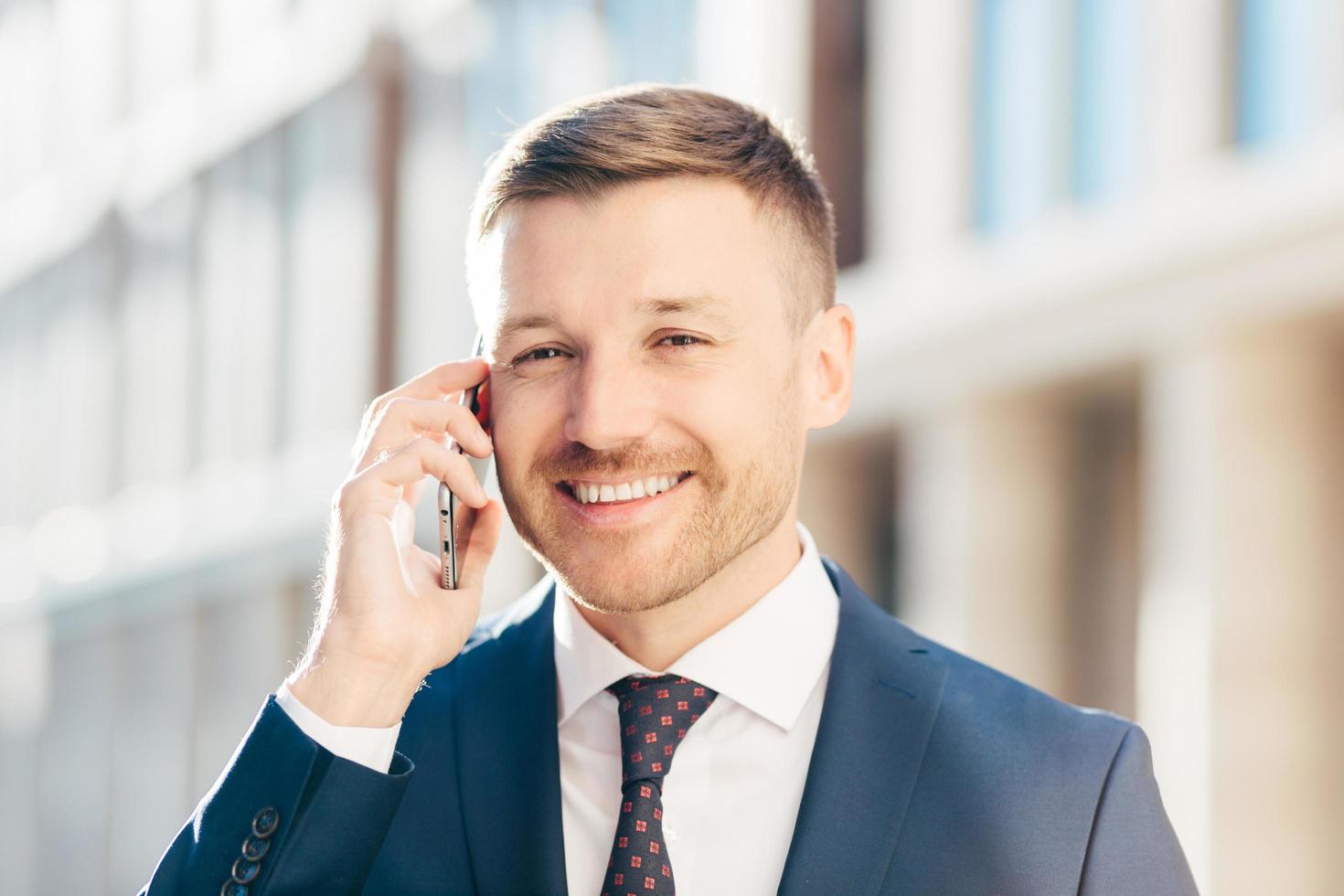 People, business and career concept. Handsome cheerful successful male in formal clothes, has bristle and positive smile, speaks via smart phone with businessman, poses against blurred background photo