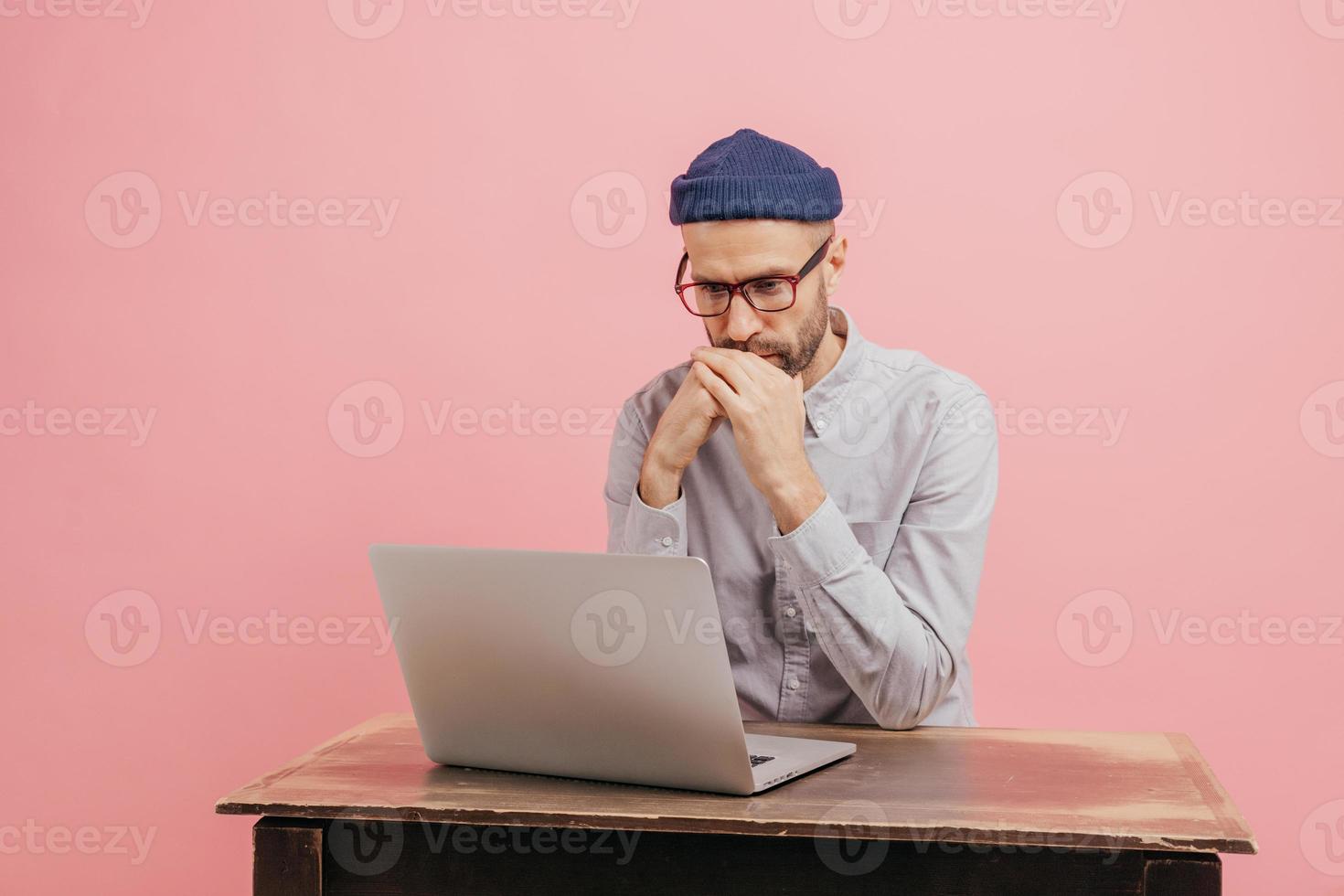 Horizontal shot of concentrated male worker reads statistics attentively, focused in laptop computer, learns something online, wears spectacles, hat and white shirt, isolated over pink wall. photo