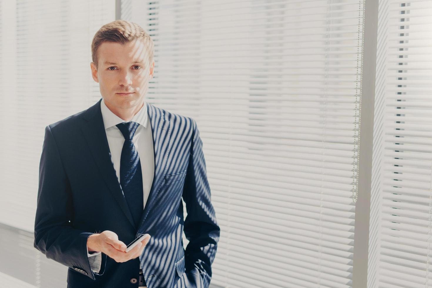 Confident male entrepreneur in formal suit, chats in social networks, recieves discount for mobile internet, has serious expression, thinks about startup, being professional worker. Technology concept photo