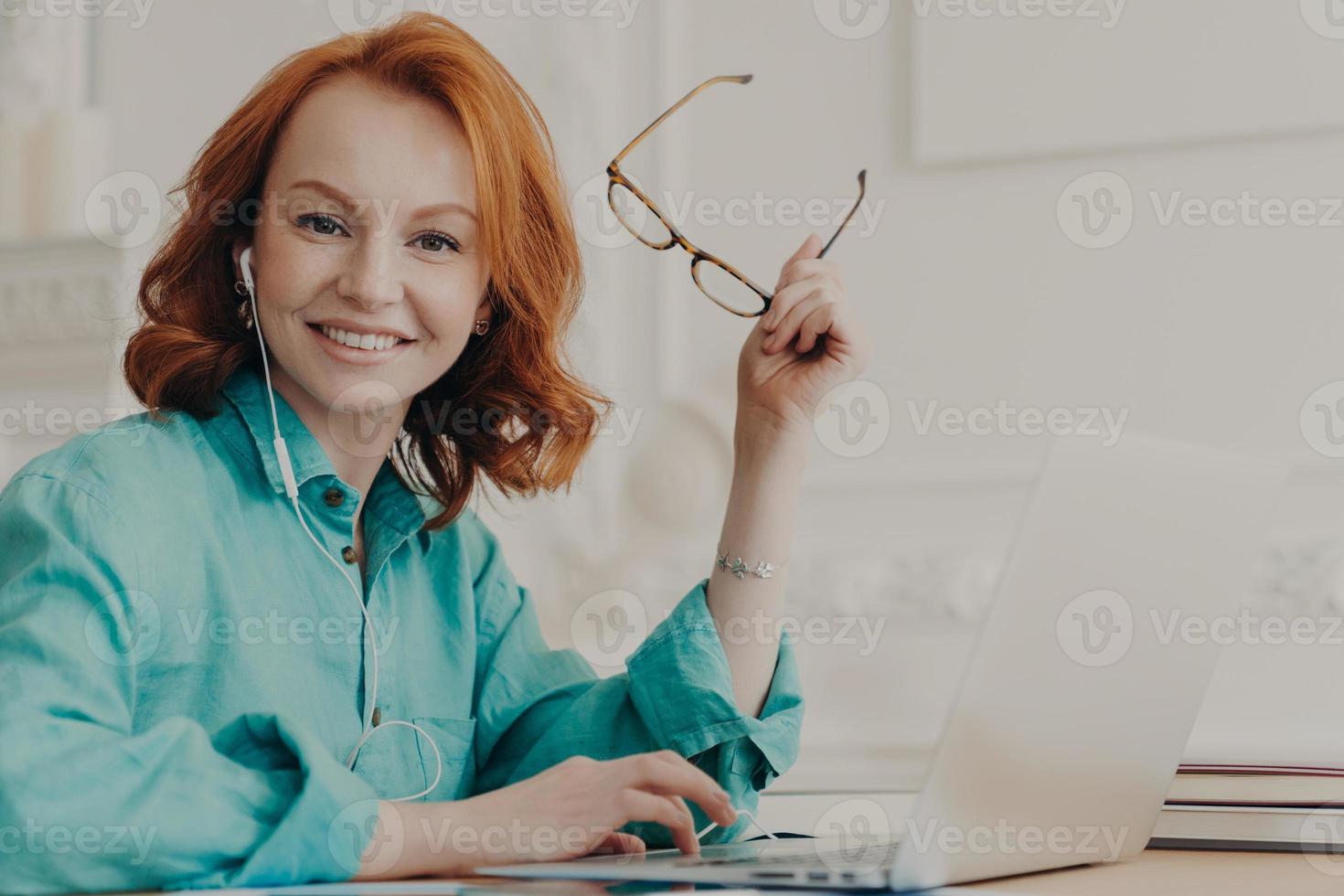 Glad red haired woman in blue shirt, works on laptop, uses earphones for watching webinar or online communication, listens courses, studies remotely from home during pandemic corona virus outbreak photo