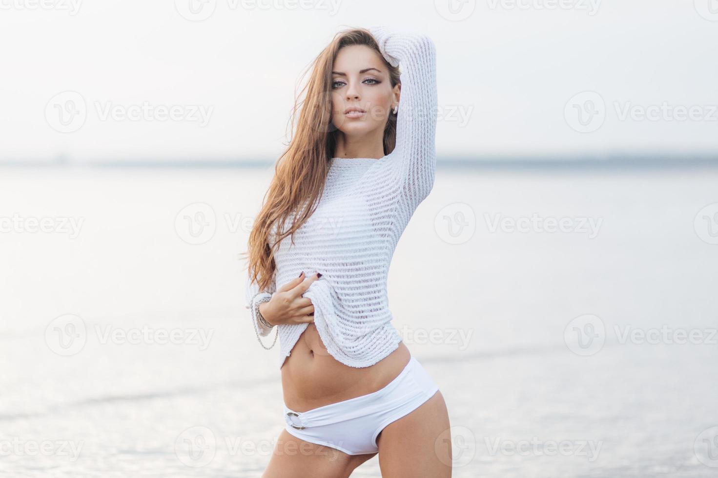 Photo of cute young woman with long hair, pleasant appearance, shows her slim belly, wears white sweater, poses against beautiful sea background. Pretty cute female models outdoor near ocean