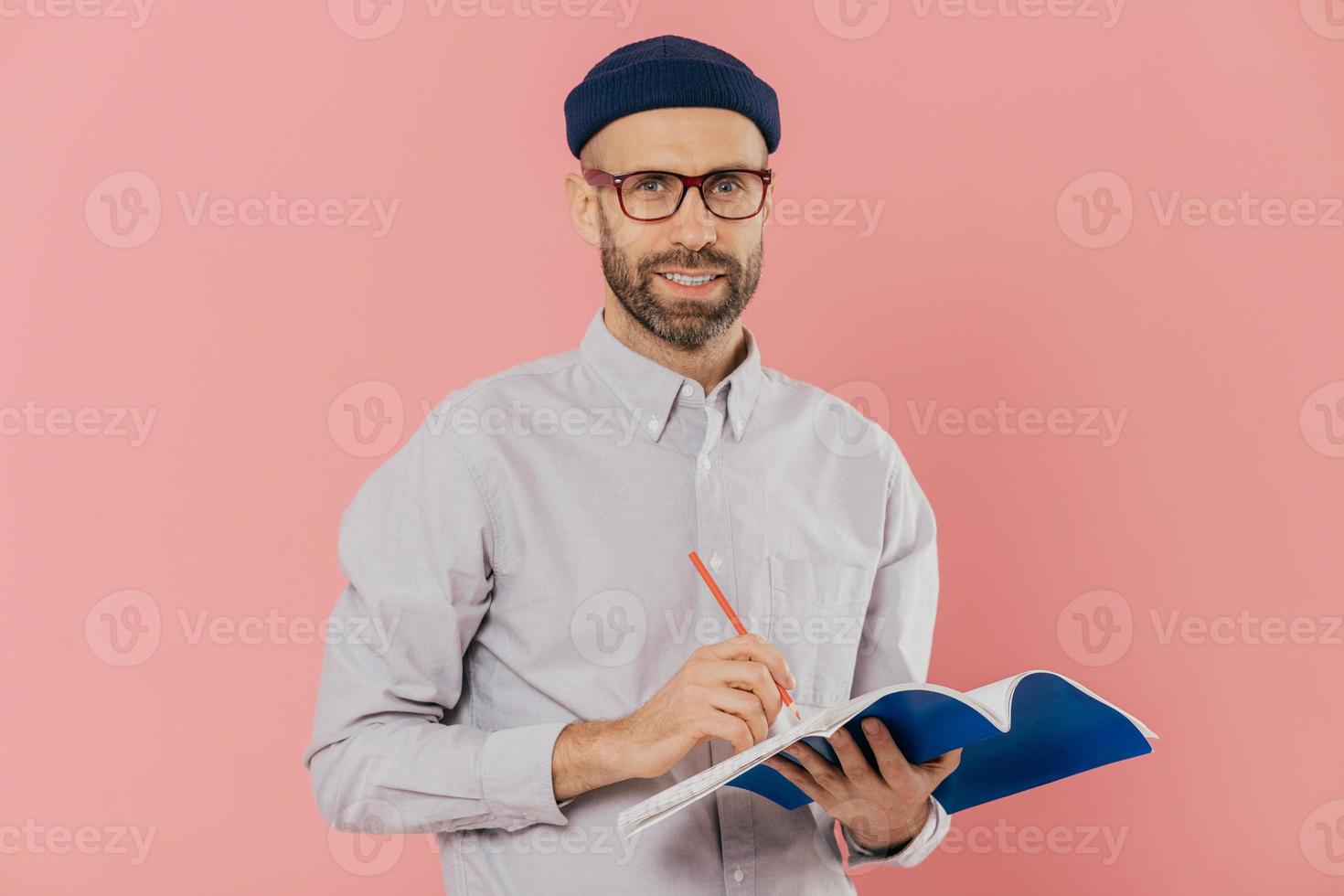 Pleased attractive man with stubble, wears transparent glasses, fashionable hat and shirt, writes with pen in notepad, prepares for speaking in front of auditorium, isolated over pink background photo