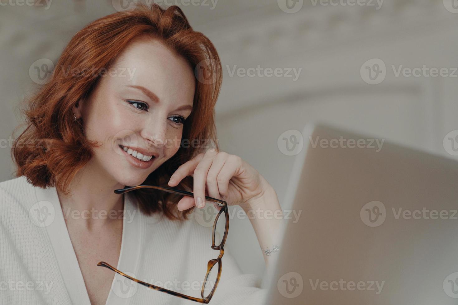 Professional smiling ginger female web designer uses application on laptop computer for creating project work, makes online research and browses internet, holds spectacles, has happy expression photo
