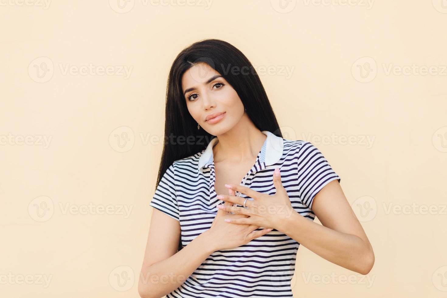 Friendly looking brunette female keeps hands on heart as expresses her gratitude, has serious expression, wears casual clothes, isolated over studio background. People, body language concept photo