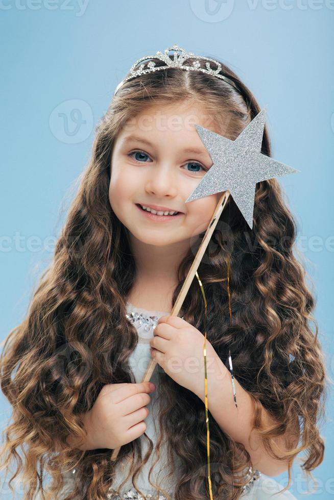 Photo of small fairy girl with curly dark hair, wears crown and dress, has blue eyes, gentle smile, holds magic wand, isolated over blue background. Angelic female kid stands indoor. Little princess