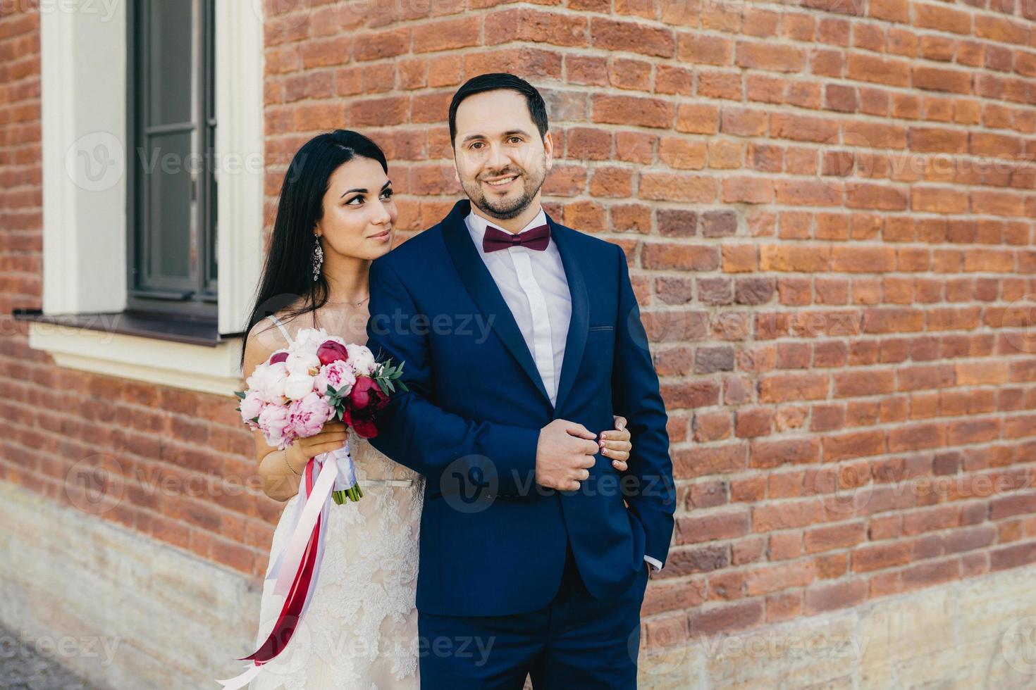 Romantic young lovely married couple posing for making photos, have happy deightful expressions. Beautiful brunette female with bouquet looks with great love at her lover, celebrate their wedding photo