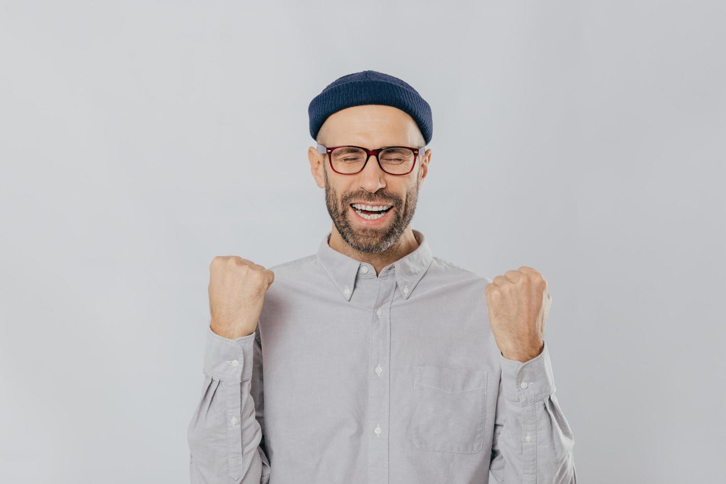 gesto de éxito. un hombre sin afeitar jubiloso y lleno de alegría levanta los puños cerrados, usa anteojos y camisa formal, celebra su triunfo, cierra los ojos de la felicidad, se siente como un verdadero ganador, aislado foto