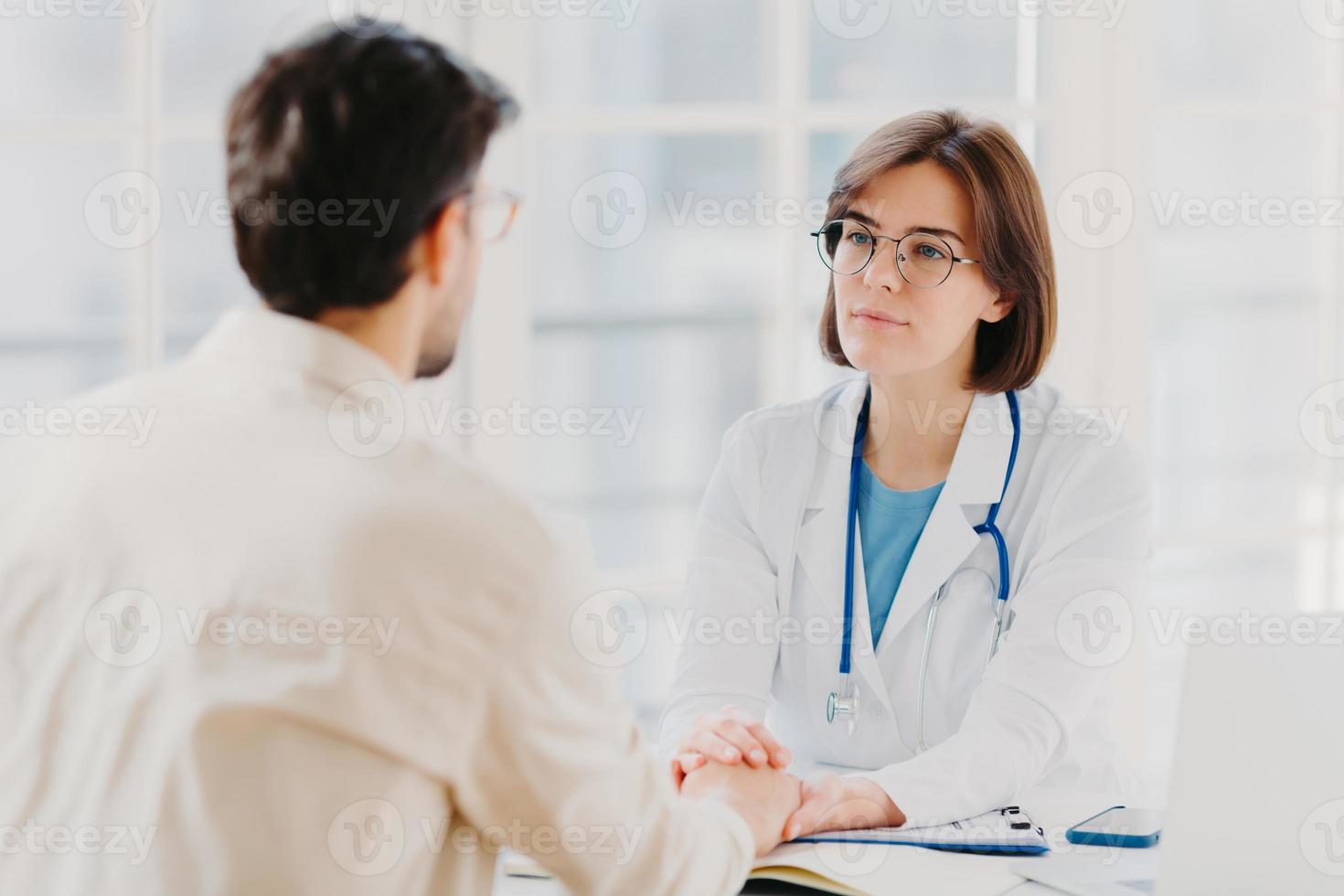 Friendly female doctor tries to support patient, holds his hands, gives useful consultation and explains medical information, makes diagnostic examining, pose in hospital room. Heathcare, assistance photo