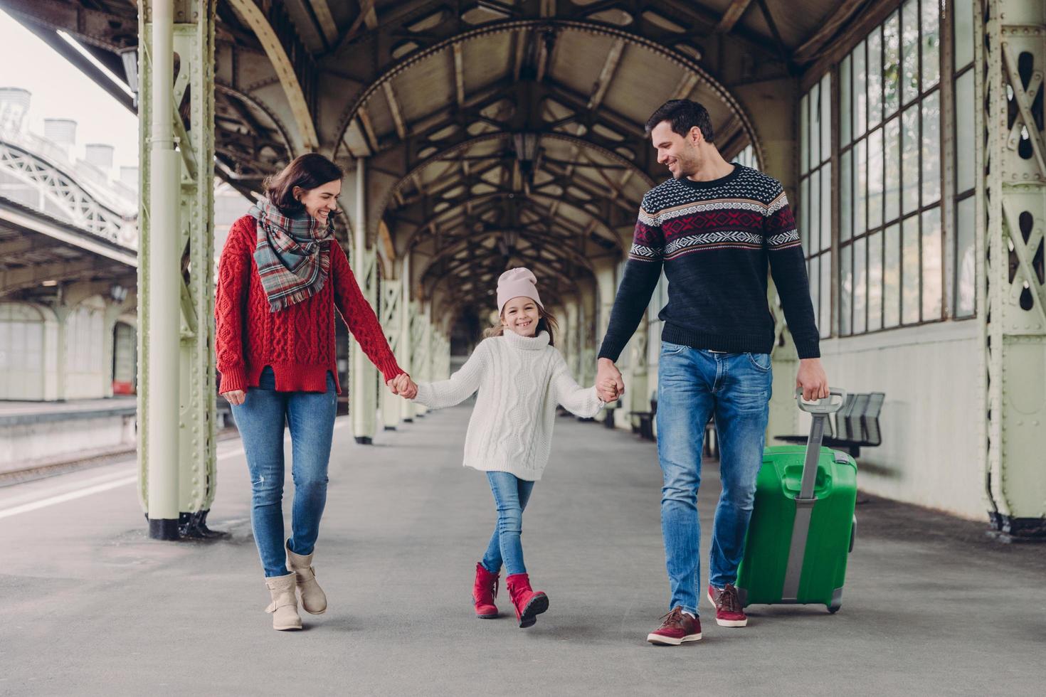 tres miembros de la familia en la estación de tren. feliz madre, hija y padre tienen expresiones faciales positivas, esperan el tren en la plataforma, disfrutan de la unión. los pasajeros contentos tienen un viaje al extranjero foto