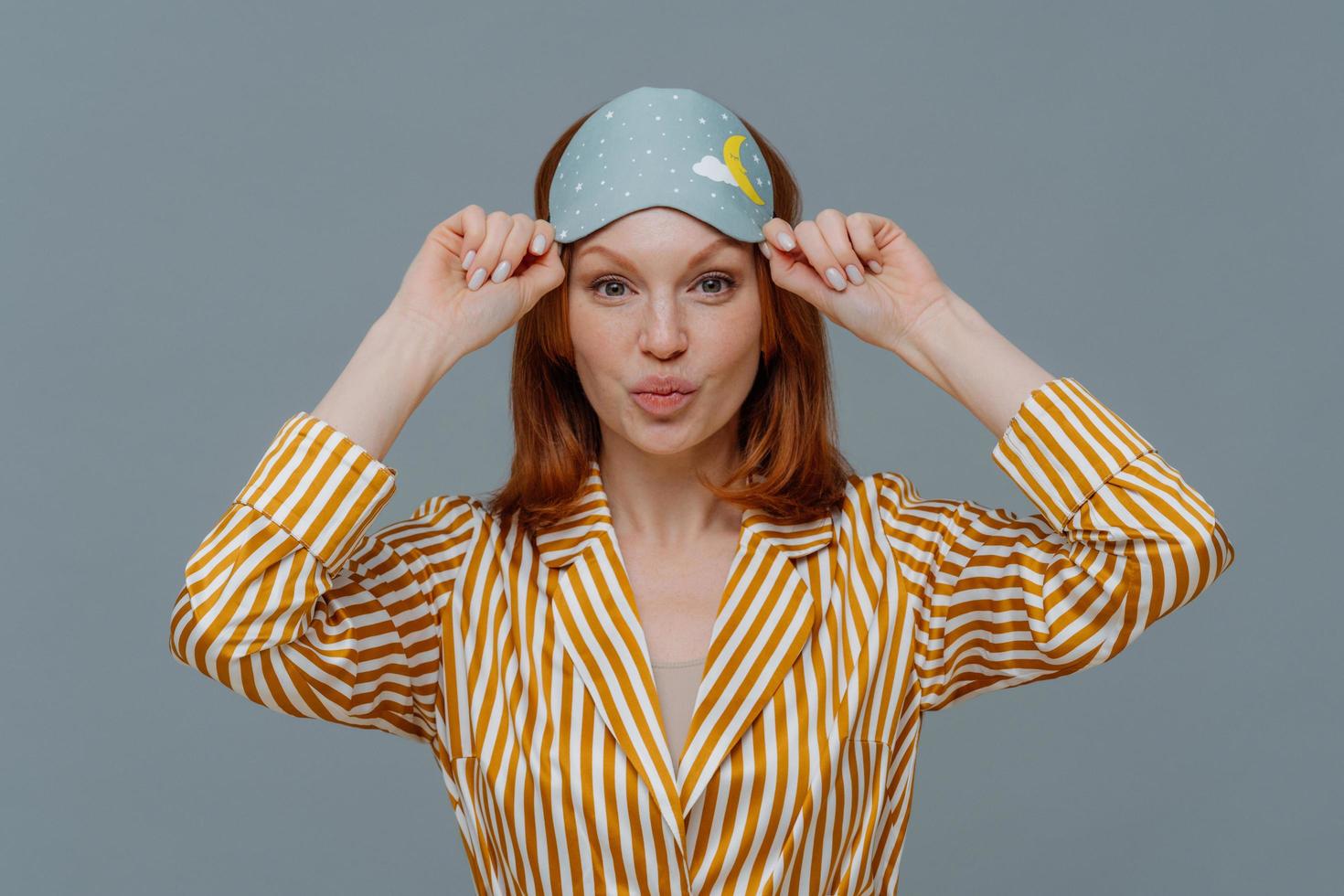 Beautiful freckled ginger woman wears sleepmask and striped pajama, keeps lips folded, has healthy skin, going to have sleep at home, enjoys day off and lazy day, isolated over grey background photo
