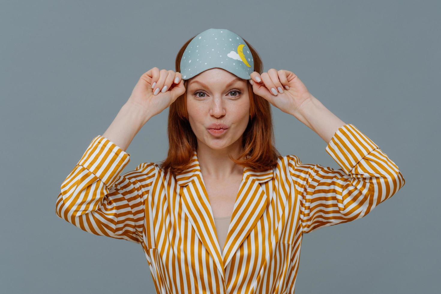 hermosa mujer pelirroja pecosa usa máscara para dormir y pijama a rayas, mantiene los labios doblados, tiene una piel sana, va a dormir en casa, disfruta el día libre y el día perezoso, aislada sobre fondo gris foto