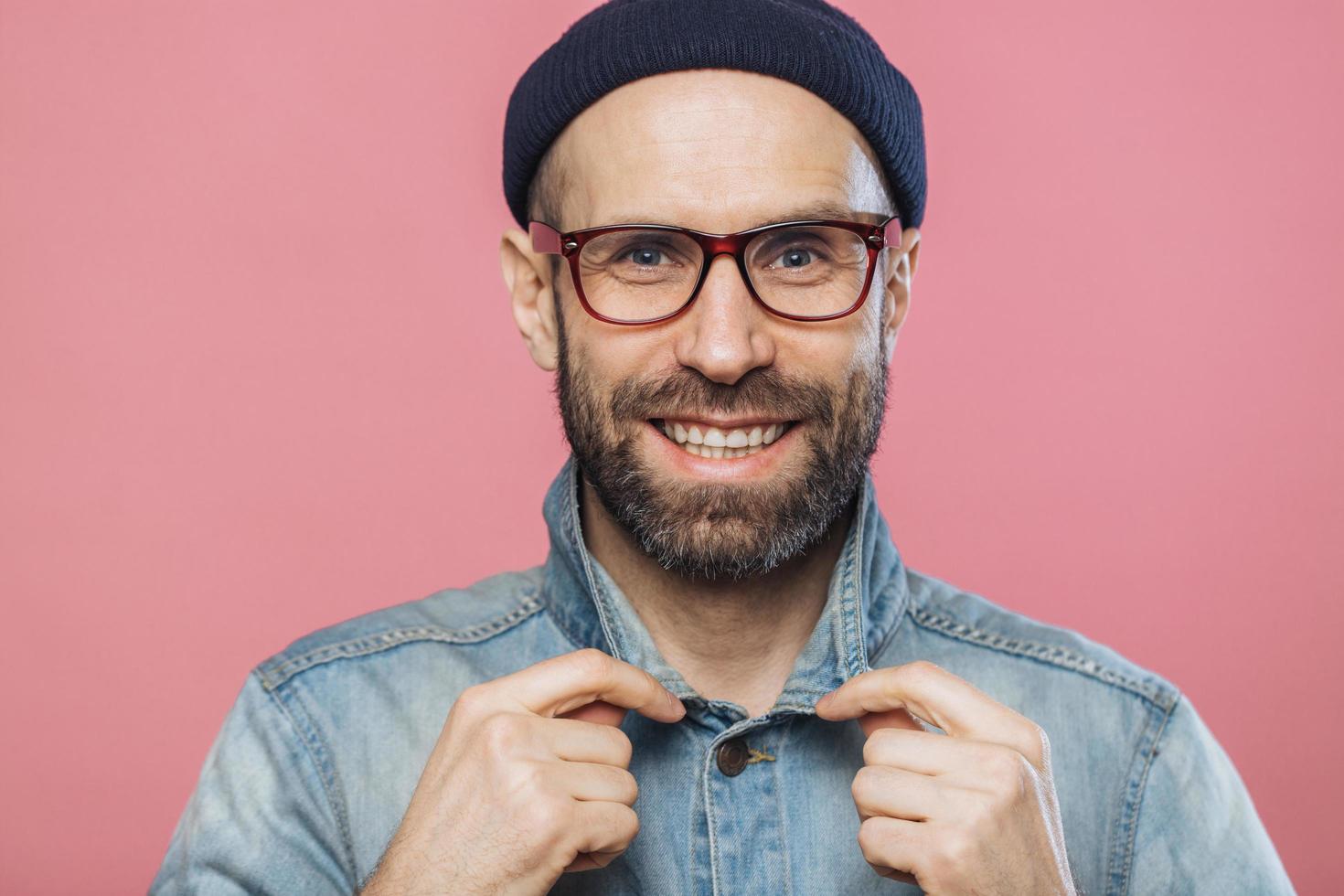 Horizontal shot of pleasant looking smiling bearded male wears glasses and denim jacket, advertizes new fashionable clothes, has happy expression, isolated over pink background. Positive emotions photo