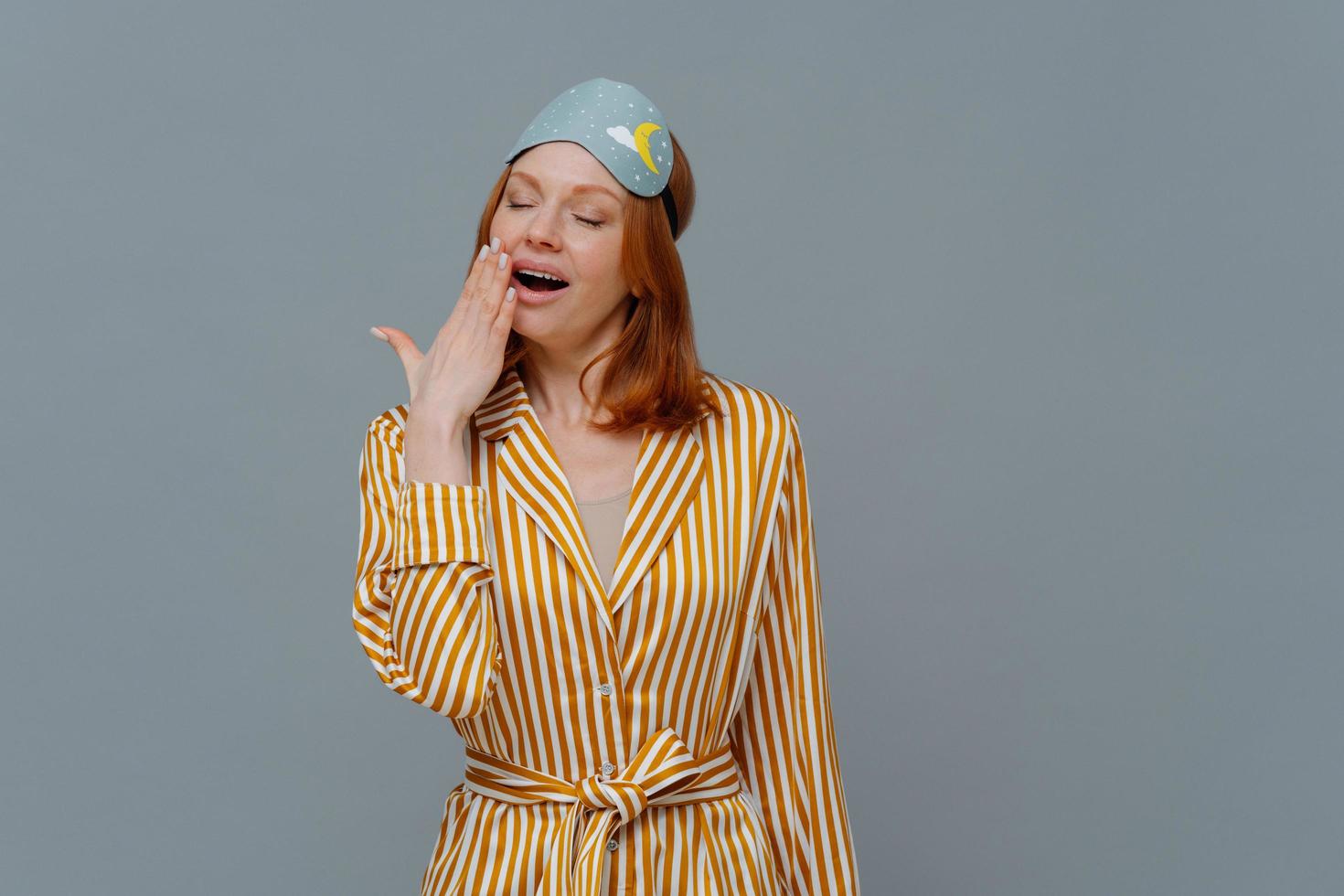 Fatigue sleepy red head woman yawns with opened mouth, wears domestic striped robe and blindfold on head, wants to sleep, stands against grey background, feels weak. Late night time concept. photo