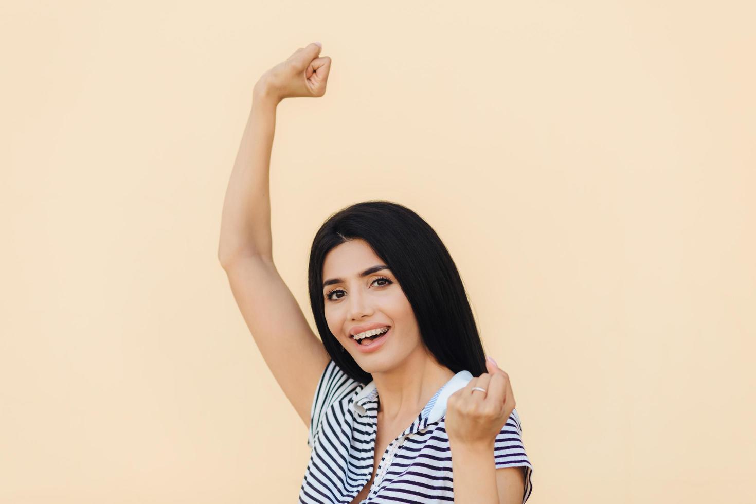 joven mujer feliz con cabello largo oscuro levanta puños con expresión alegre, tiene un aspecto atractivo, usa ropa informal, se regocija de su éxito, tiene triunfo. concepto de personas, logros y felicidad foto