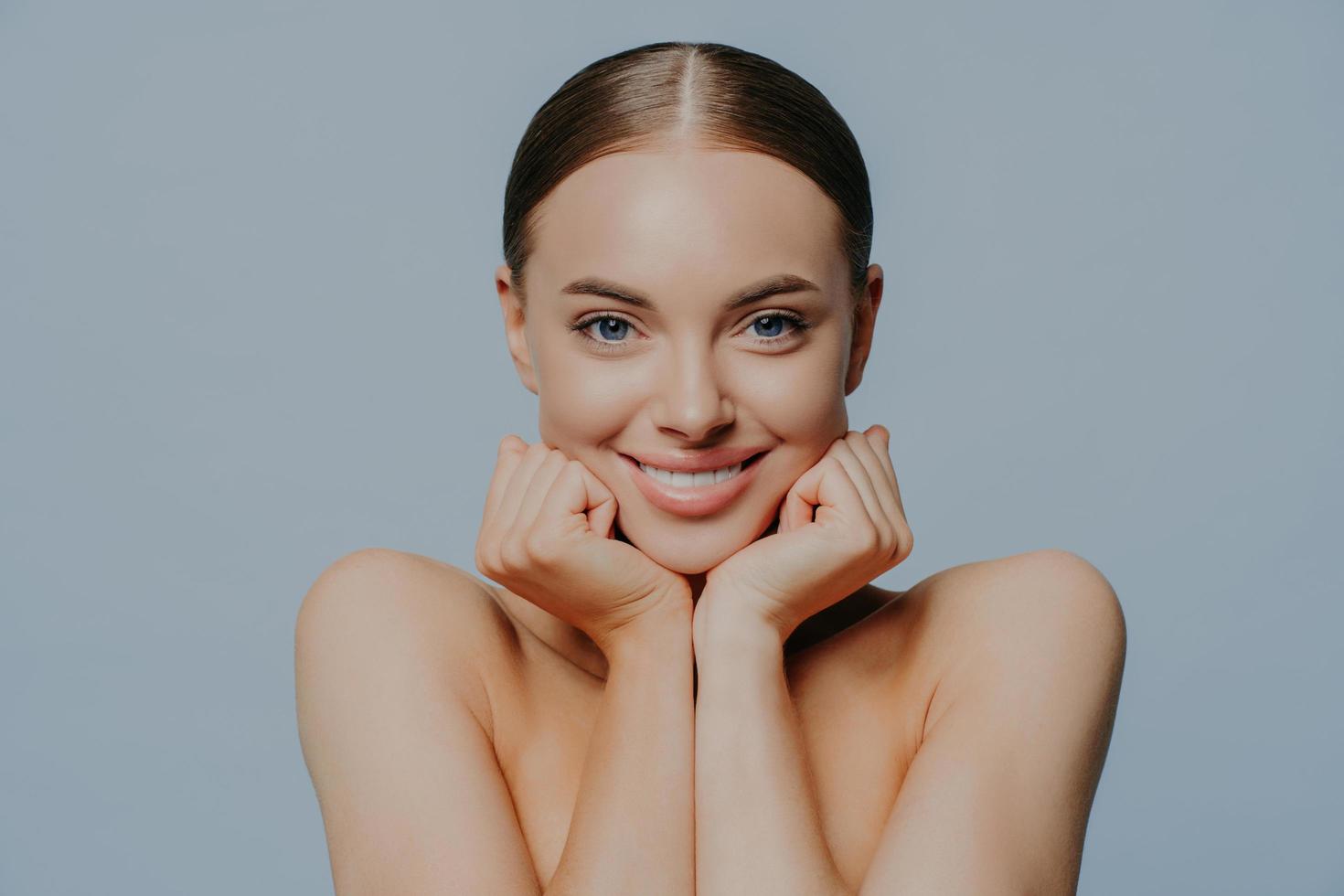 Women, skin care and tenderness concept. Young female takes care of skin and body health, keeps hands under chin and smiles gently, wears makeup, has combed hair, delighted with skin care product photo