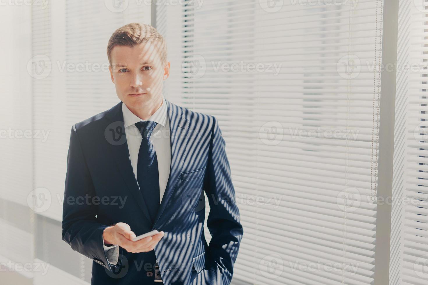 Candid shot of prosperous businessman checks notification on cellular, plans daily schedule, wears formal suit, stands near window, looks directly into camera, has work break. Office routine photo