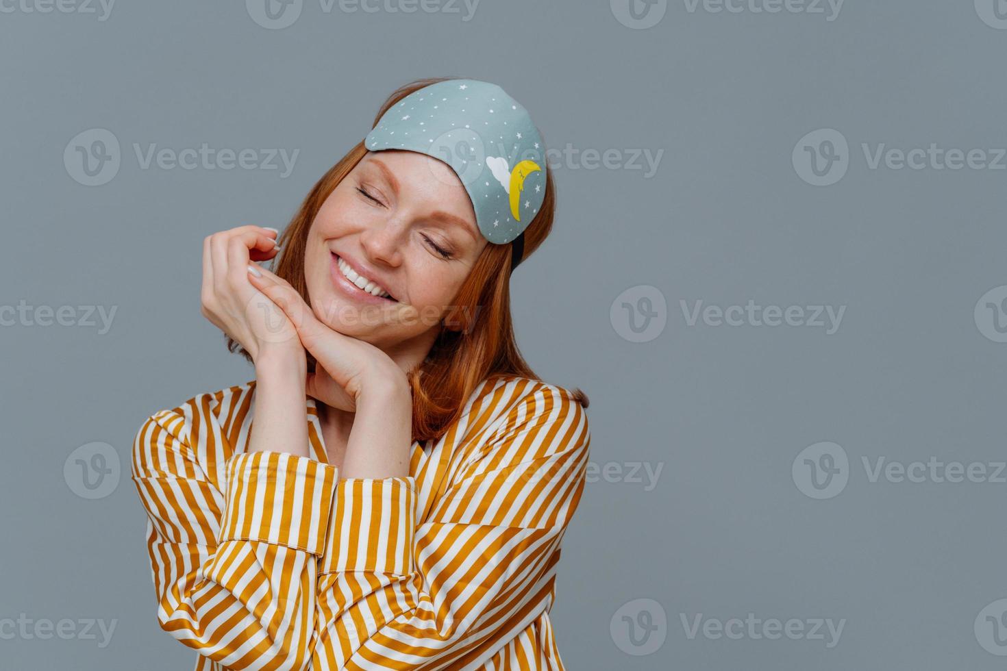 retrato de una mujer alegre y de buen aspecto inclina la cabeza y sonríe suavemente, imagina algo agradable, usa máscara para dormir o venda en los ojos y pijama, aislada sobre fondo gris, posa en un dormitorio acogedor foto