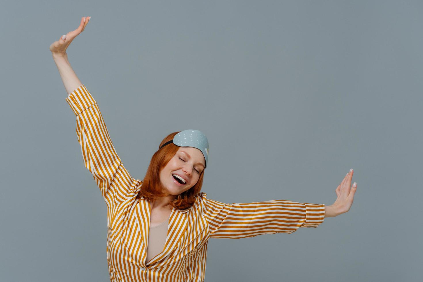 Good morning and awakening concept. Happy relaxed red haired young woman wears comfortable soft pajamas and sleepmask, stretches arms sideways, has glad expression, isolated on grey background. photo