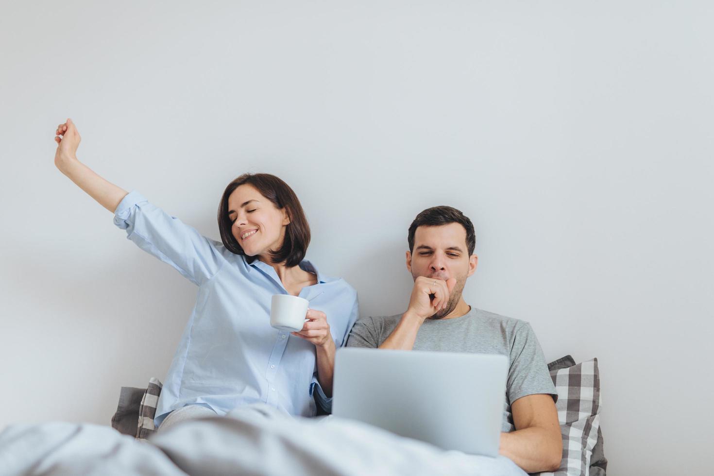 Glad female stretches in bed, drinks hot coffee and her husband works all night on laptop computer, yawns as wants to sleep, spend time together in bedroom, isolated over white background photo