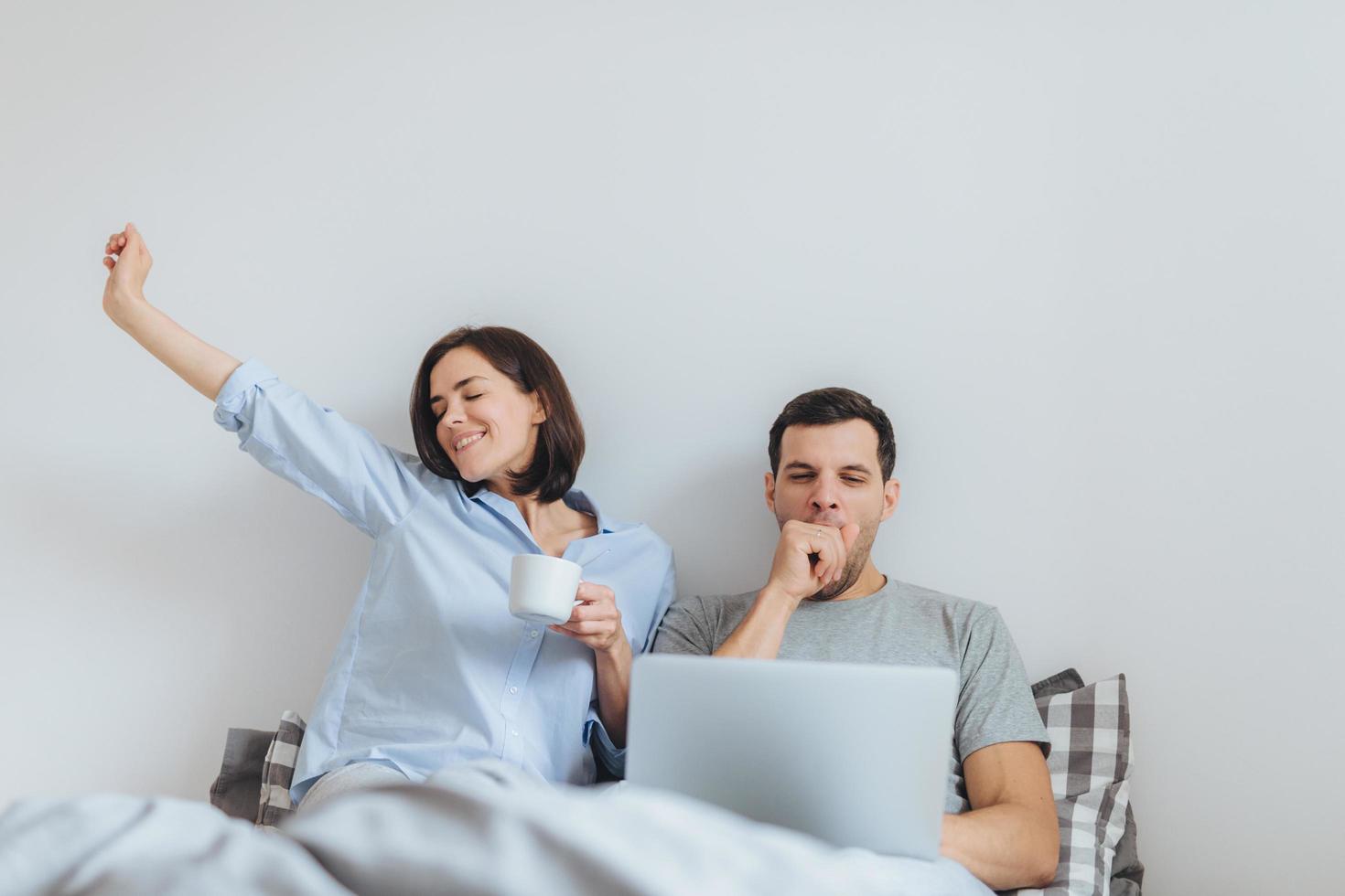 la mujer feliz se estira en la cama, bebe café caliente y su esposo trabaja toda la noche en una computadora portátil, bosteza como quiere dormir, pasan tiempo juntos en el dormitorio, aislados sobre fondo blanco foto