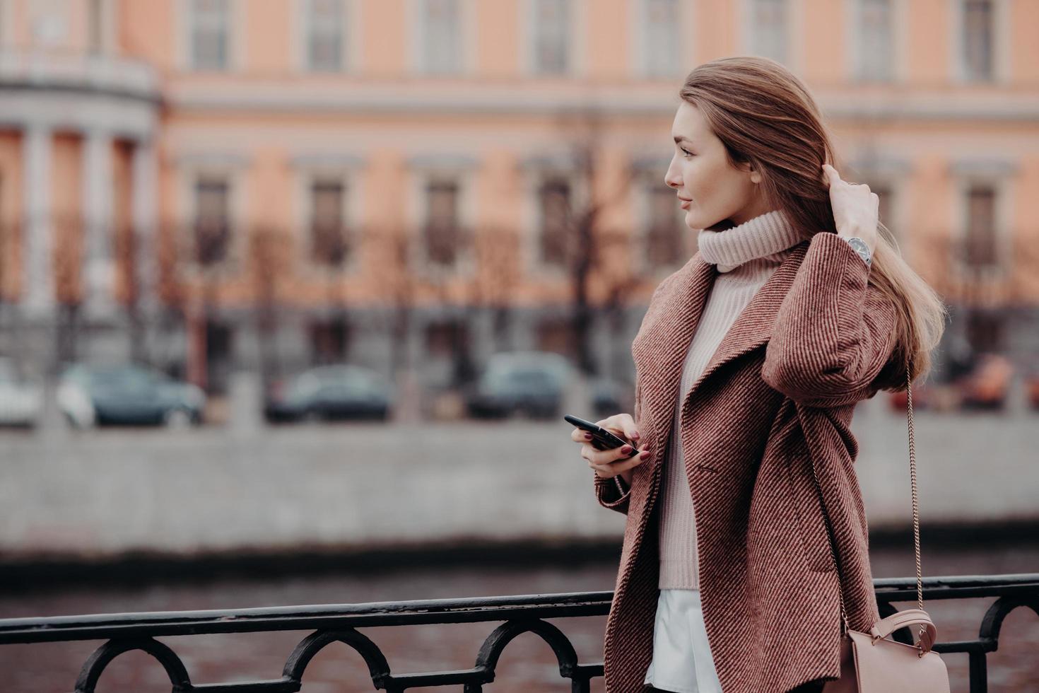 Outdoor shot of relaxed young female looks aside with thoughtful expression, holds modern cell phone, waits for call, dressed in white sweater and coat, carries bag, stand on bridge in big city photo